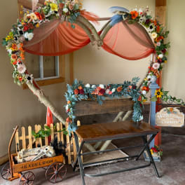 Floral arch with orange drapes and mixed flowers around a rustic bench.