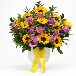 Bouquet of yellow sunflowers and lavender roses in a white vase with a yellow ribbon