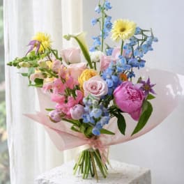 Pastel bouquet with roses, peonies, calla lily, and blue flowers wrapped in pink paper