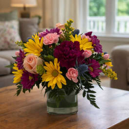 Mixed bouquet of yellow daisies, pink roses, and purple flowers in a glass vase
