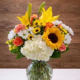 Mixed bouquet of yellow lilies, sunflowers, roses, daisies, and hydrangea in a glass vase