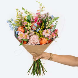 Handheld bouquet of mixed pink, peach, blue, and yellow flowers wrapped in brown paper