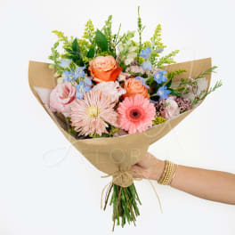 Hand-tied bouquet of pink, peach, and blue flowers wrapped in kraft paper