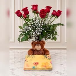 Red roses arranged above a teddy bear and floral gift box