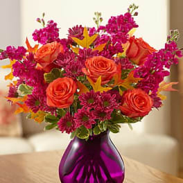 Bright bouquet of orange roses and magenta flowers in a purple glass vase on a wooden table