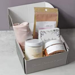 Open gray gift box with spa items including jars, boxed products, and a pink fabric accessory.