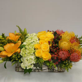 Long floral centerpiece with orange lilies, yellow roses, and mixed blooms in a rectangular vase