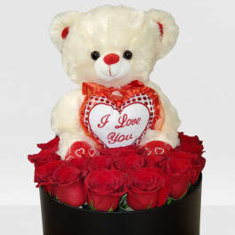 Cream teddy bear holding an 'I Love You' heart atop a round box of red roses