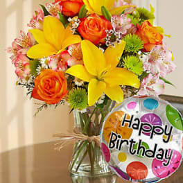 Bouquet of yellow lilies, orange roses, and pink flowers in a glass vase with a birthday balloon