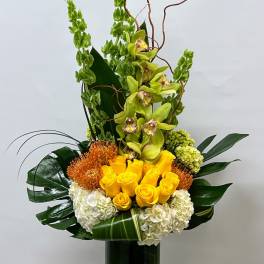 Tall floral arrangement with yellow roses, green orchids, and white hydrangeas in a glass vase