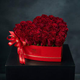 Red roses arranged in a red heart-shaped box with a ribbon bow