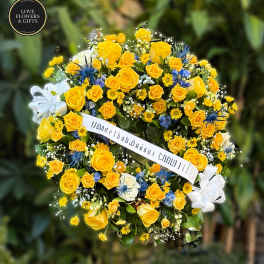 Round standing wreath of yellow roses and daisies with blue accents and a white ribbon banner.