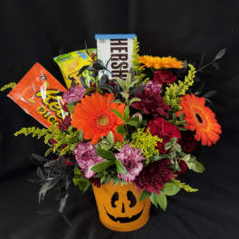 Orange and burgundy flower arrangement in a jack-o'-lantern pot with candy bars