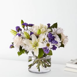 White lilies and daisies with purple filler in a clear glass vase