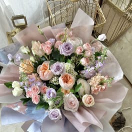 Pastel bouquet of pink, peach, and lavender roses with white accents wrapped in soft mauve paper