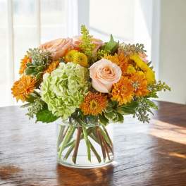 Round arrangement of peach roses, green hydrangea, and orange mums in a clear glass vase