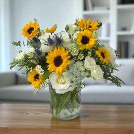Yellow sunflowers and white roses arranged with small daisies in a clear glass vase on a wooden table