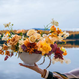 Low centerpiece of yellow, peach, and cream flowers in a white pedestal bowl held by a hand.
