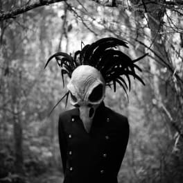 Person in a dark coat wearing a skull-like mask and feathered headpiece in a wooded setting