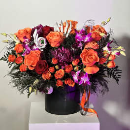 Orange and purple floral arrangement with roses, orchids, and Halloween skeleton hand accents in a black container