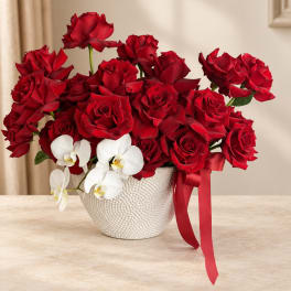Red roses in a white textured vase with white orchids and a red ribbon