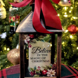 Decorative lantern with a red bow and Christmas message in front of a lit tree