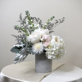 Asymmetrical arrangement of white and blush flowers in a gray cylinder vase on a draped ivory table.