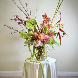 Tall modern arrangement with pink, green, and orange flowers in a glass vase on a draped round table