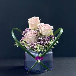 Three pale pink roses in a purple glass vase with green ribbon accents