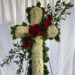 Tall white floral cross with red roses and greenery displayed on a metal easel