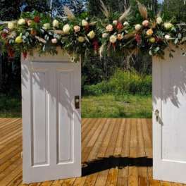 Tall rustic doorway arch made of two white doors topped with a garland of roses and grasses