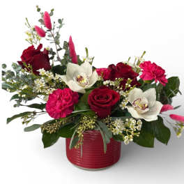 Low arrangement of red roses, hot pink carnations, and white orchids in a red ceramic vase