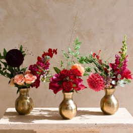 Three floral arrangements in gold vases with red, pink, and peach blooms