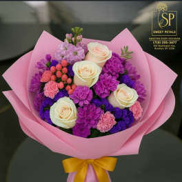 Pink-wrapped bouquet of cream roses, purple blooms, and pink accents with a yellow ribbon