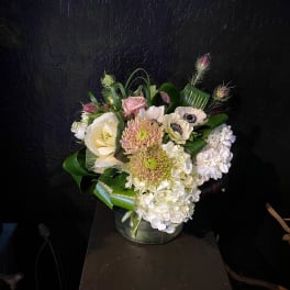 Compact pastel flower arrangement with hydrangeas, mums, anemones, and roses in a clear glass vase.