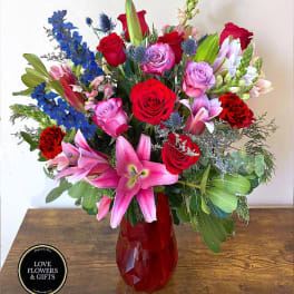 Tall arrangement of red roses, pink lilies, purple roses, and blue delphinium in a red glass vase.