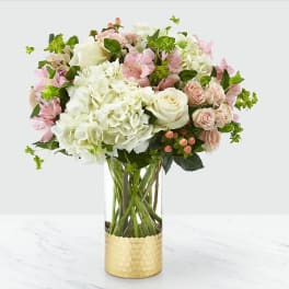 Round arrangement of white hydrangeas, white roses, and soft pink blooms in a clear glass vase with a gold base