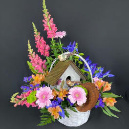 Basket arrangement with pink snapdragons, gerbera daisies, blue irises and a birdhouse centerpiece