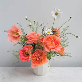 Peach roses and poppy-style blooms with white daisies in a small white ceramic vase