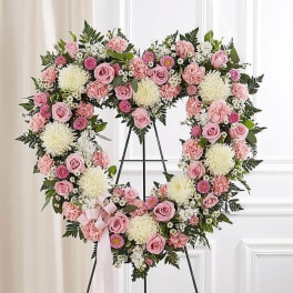 Heart-shaped standing wreath of pink roses, carnations, and white mums with a bow on a metal easel