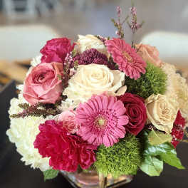 Round arrangement of pink gerbera, roses, carnations, and cream blooms in a glass cube vase