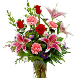 Tall glass vase with red roses, pink lilies, pink carnations, and purple accent flowers