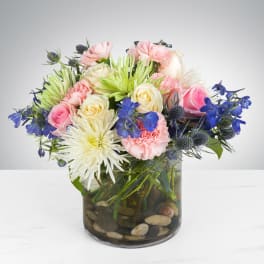 Round bouquet of pink roses and carnations with white mums and blue flowers in a glass vase with stones