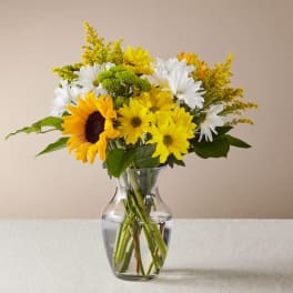 Clear glass vase of yellow daisies, a sunflower, white daisies, and other mixed flowers.