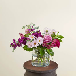 Low pink and purple mixed flower arrangement in a clear glass vase on a round stand