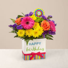 Bright birthday arrangement with pink gerbera, orange roses, yellow mums in a happy birthday cube vase