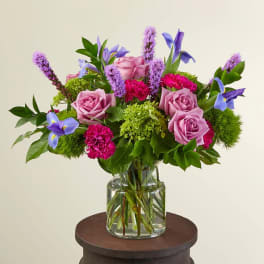 Mixed bouquet of lavender roses, bright pink carnations, blue iris and green blooms in a clear glass vase.