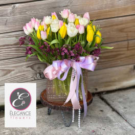 Pastel pink, white, and yellow tulips with purple accents in a glass vase with pink ribbon and pearls.