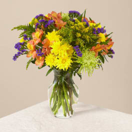 Mixed bouquet of yellow and green mums, orange alstroemeria, and purple flowers in a clear glass vase