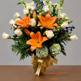 Tall arrangement of orange lilies, white roses, blue flowers, and hydrangeas in a gold vase with a bow